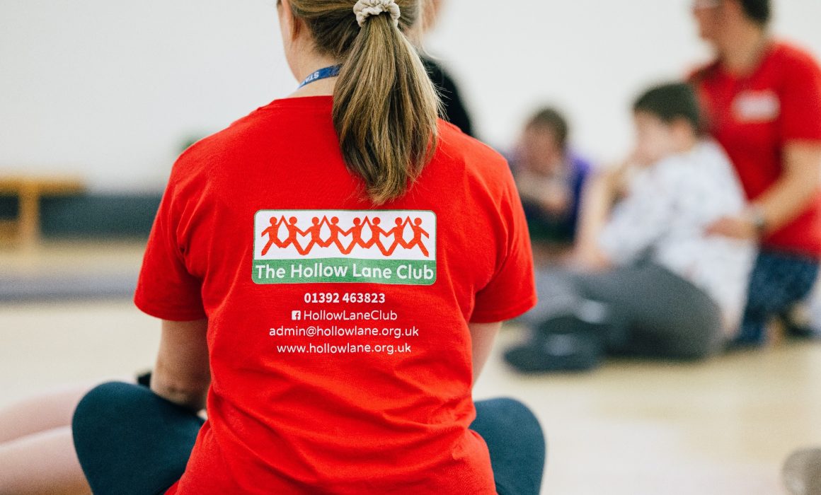 A support worker in a red Hollow Lane Club tshirt with her back to the camera. She is sat cross legged on the floor and there are other people in the background taking part in the club.